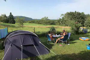 Campingzelt auf grünem Rasen, daneben Campingstühle und Tisch im Garten, im Hintergrund Bäume und Hügel.