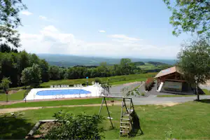 Blick vom Campingplatz ins Tal: Pool, Rasenfläche, Weg und ein Holz-Pavillon am Hang mit Bäumen und Hügeln.