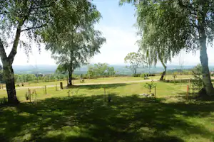 Grünes Campinggelände mit Rasen, Bäumen und eingezäunten Parzellen, dahinter weite Landschaft und Hügel.