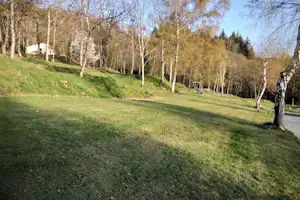 Grüne Campingwiese mit Bäumen im Wald, ein Weg führt am Hang entlang, im Hintergrund ein kleines Gebäude.