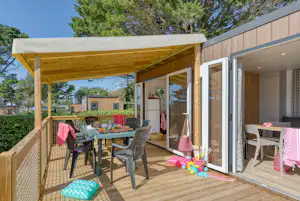 Holz-Terrasse am Mobilheim mit Esstisch, Stühlen und Spielzeug, daneben große Schiebetür und Fensterfront, umgeben von Bäumen.