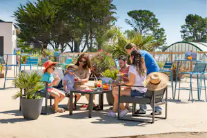 Familie sitzt an einem Camping-Tisch mit Getränken und Sonnenschirm, im Hintergrund Pool und Bäume.