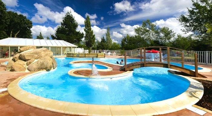 Großes Camping-Schwimmbecken mit rundem Whirlpool, Sonnendeck und Zaun, im Hintergrund Bäume und ein Gebäude mit Glasdach.