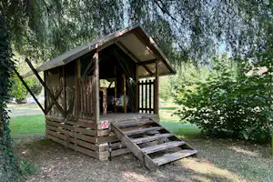 Holzhütte auf einer Holzplattform mit Vorhang, unter Bäumen im Campinggelände, mit Sitzbank und Nummernschild L9.