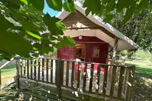 Zelt mit roter Außenwand und Veranda, umgeben von grünen Blättern und Holzgeländer auf dem Campingplatz.