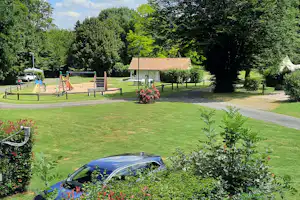 Kleines Haus mit Satteldach auf dem Campingplatz, daneben Spielplatz, Bänke und Tische im grünen Garten.