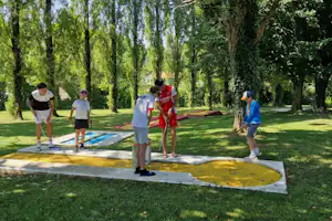 Minigolf im Campingpark: Kinder und Erwachsene stehen an einem Bahnfeld auf dem Rasen zwischen Baeumen.