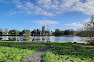 Seeufer mit Kiesweg im Park, grünes Gras, Bäume und hohe Pappeln am Wasser unter blauem Himmel mit Wolken.