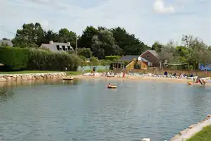 Blick auf den See mit Schwimmzone, Ruderbooten und Liegewiese am Campingplatz, im Hintergrund Bungalows und Bäume.