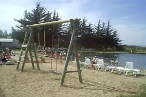 Spielplatz mit Schaukel und Sandfläche am See, daneben Liegestühle und Bäume am Campingplatz.