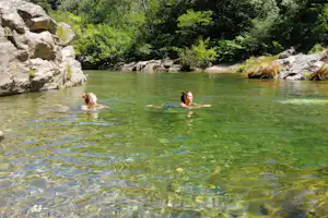 Zwei Personen schwimmen im klaren Fluss, umgeben von Felsen und dichtem Grünwald.