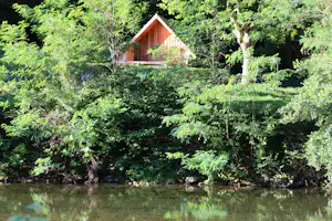 Holzhütte mit Terrasse im Grünen am Fluss, umgeben von Bäumen und Sträuchern, Spiegelung im Wasser.
