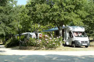 Wohnmobil neben Blumenbeet und Zeltvorbau unter Bäumen auf einem Campingplatz, mit gepflastertem Stellplatz.