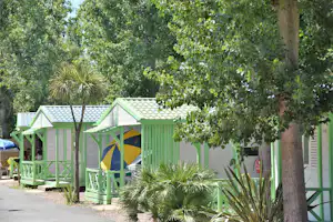 Grünes Holzhaus mit überdachtem Eingang und Sitzbereich, daneben ein Zelt auf dem Campingplatz, umgeben von Bäumen und Sträuchern.