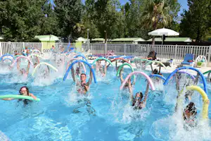 Kinder im Camping-Schwimmbecken mit bunten Wasserreifen, Spritzwasser und Sonnenschirm, im Hintergrund Bäume und Zelte.