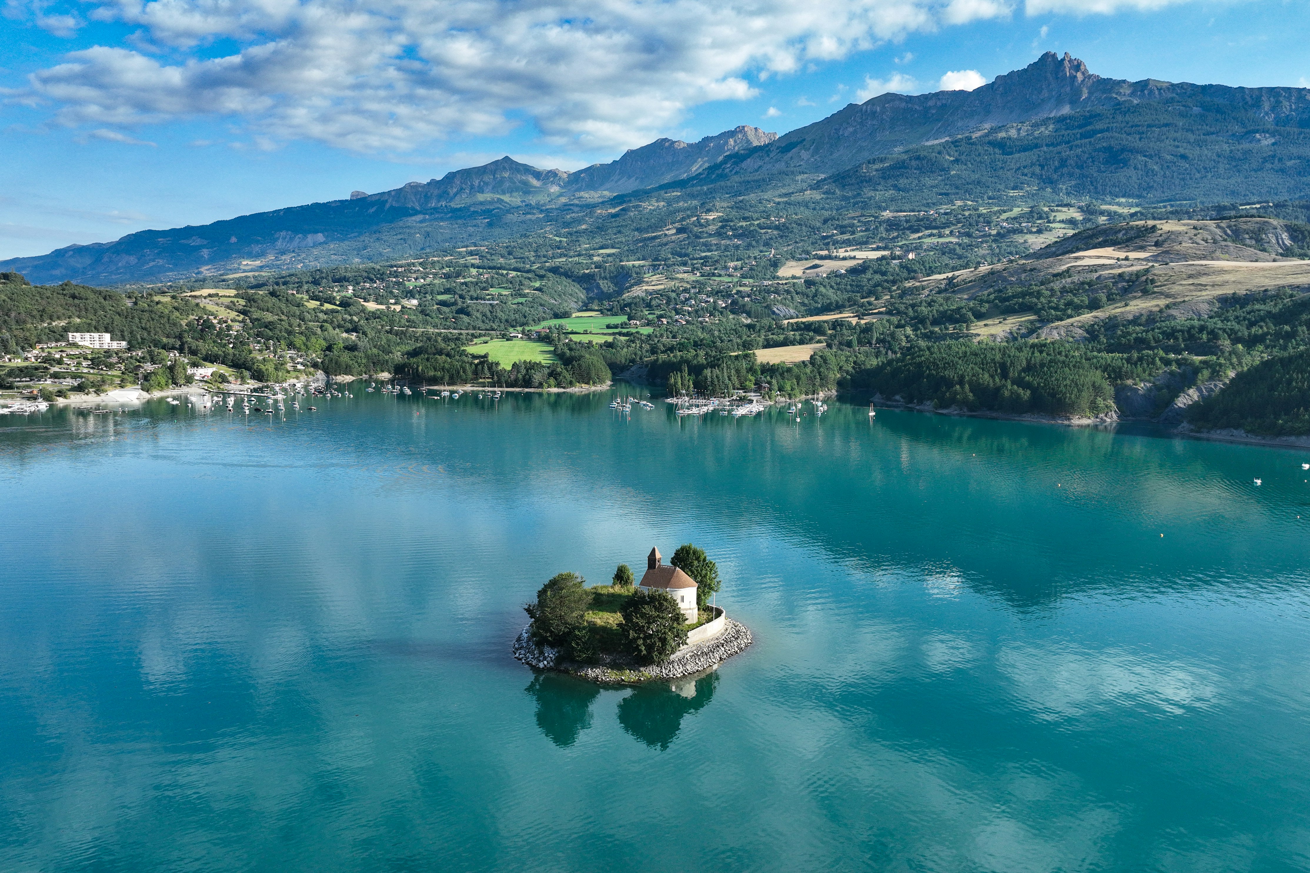 Kleines Inselchen mit Baum und Felsen im ruhigen See, dahinter grüne Berge und ein Dorf am Ufer.
