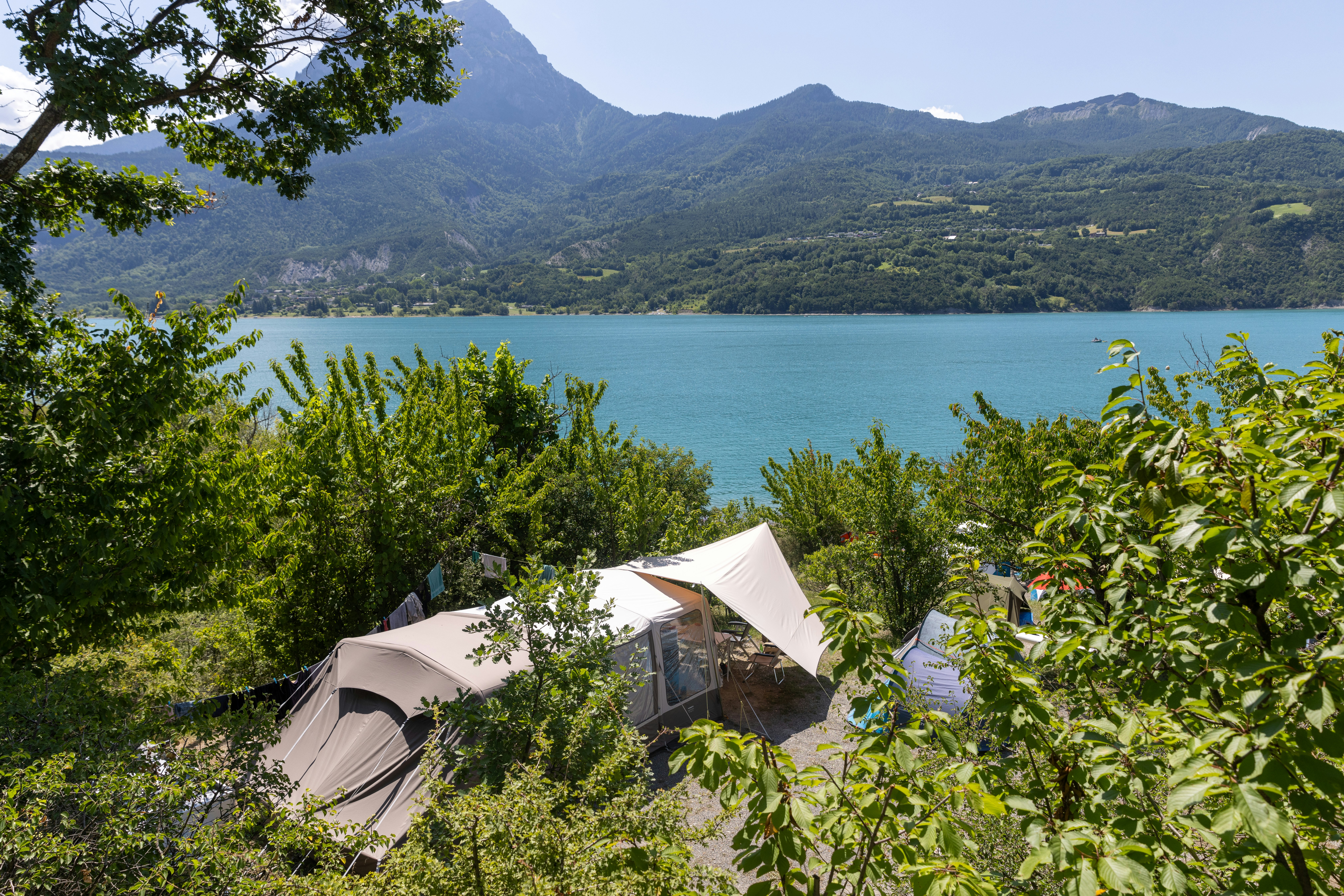 Zelt auf einem Campingplatz mit Blick auf den See, umgeben von Büschen und Bäumen, im Hintergrund Berge.