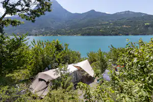 Zelt auf einem Campingplatz mit Blick auf den See, umgeben von Büschen und Bäumen, im Hintergrund Berge.