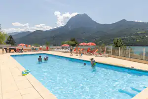 Blick auf den Camping-Pool mit Liegen, Personen im Wasser und roten Sonnenschirmen, im Hintergrund Berge und grünes Tal.
