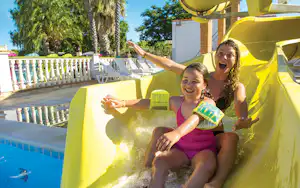 Zwei Kinder sitzen auf einer gelben Poolrutsche am Campingplatz, im Hintergrund weiße Liegen und Bäume.