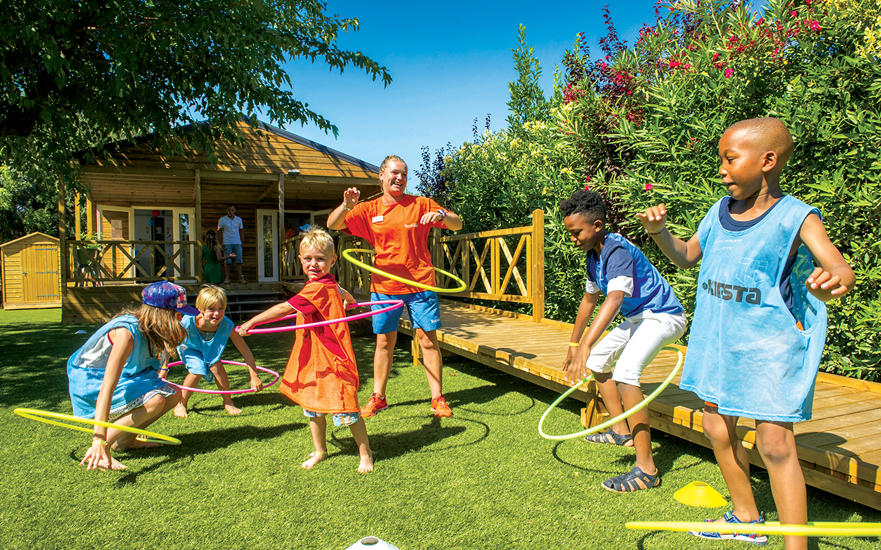 Kinder und Erwachsene spielen im Campinggarten auf Rasen, mit Spielgeräten und Holzhäuschen im Hintergrund.