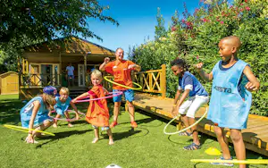 Kinder und Erwachsene spielen im Campinggarten auf Rasen, mit Spielgeräten und Holzhäuschen im Hintergrund.