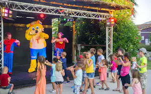 Kinder und Familien stehen vor einer Bühne beim Campingfest, ein Maskottchen und Animateure sind sichtbar.