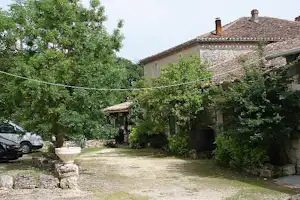 Haus mit Steindach und Eingang im Campinghof, umgeben von Bäumen, mit Sitzbereich und geparktem Auto links.
