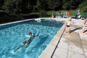 Camping-Pool mit klarem Wasser, Steinsteg und Liegen; im Hintergrund Bäume und ein paar Sonnenschirme.