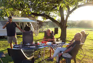 Camping-Tisch mit Grill und Essen, daneben Wohnmobil und Stühle im Schatten eines Baums auf einer Wiese am See.