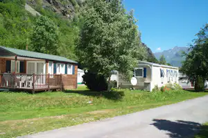 Reihe von Mobilheimen am Campingplatz mit Holzterrasse, Bäumen und Zufahrtsstraße, im Hintergrund Berge und Tal.