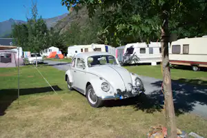 Klassischer Volkswagen Oldtimer auf einem Campingplatz, daneben Wohnwagen und Zelte, Rasenfläche und Bäume im Hintergrund.
