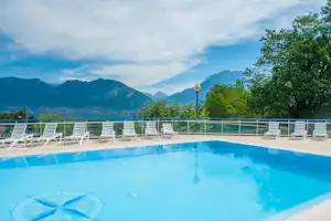 Campingpool mit Liegestühlen am Beckenrand, Blick auf Berge und Bäume bei bewölktem Himmel.