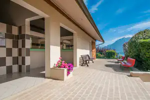 Terrasse am Campinggebäude mit Bank, roten Stühlen, Blumenbeet und Blick auf Berge im Hintergrund.