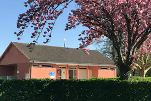 Campinggebäude mit Satteldach und rosa blühendem Baum im Vordergrund, Hecke und Himmel im Hintergrund.