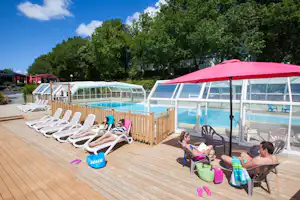 Holzdeck am Campingplatz mit Sonnenliegen, Kindern und einem roten Sonnenschirm neben dem beheizten Pool im Freien.