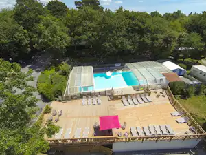 Schwimmbecken mit Glasabdeckung und Sonnenliegen auf dem Campingplatz, umgeben von Bäumen und einem Holzdeck.