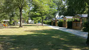 Grüner Campingplatz mit Rasenfläche, gepflastertem Weg und Bäumen; im Hintergrund stehen kleine Gebäude und Unterstände.