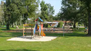 Spielplatz mit Klettergerüst und Rutsche auf einer Wiese, im Hintergrund Bäume und Campinggebäude.