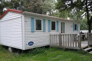 Wohnwagen mit Veranda, blauen Fensterläden und großen Fenstern, umgeben von Bäumen und Rasen am Campingplatz.