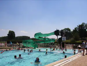 Grüne Wasserrutschen über dem Pool, Kinder planschen im Wasser, daneben Zaun und Bäume am Campingplatz.