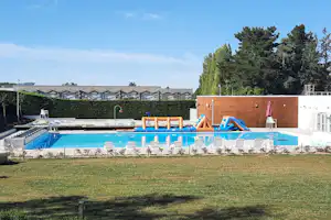 Schwimmbad mit blauem Wasser, Liegen und Sonnenschirmen vor einem Holz- und Gebäudebereich, umgeben von Rasen und Bäumen.