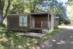 Holzbungalow mit Veranda, Stufen und Sitzbereich vor dem Eingang, umgeben von Bäumen und Rasen.