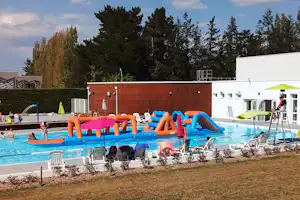 Aufblasbares Wasser-Spielgerät mit Rutschen im Camping-Schwimmbecken, daneben Liegen und ein weißes Gebäude mit Sonnenschirm.