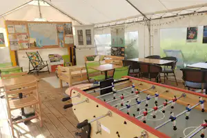 Spielbereich im Campingzelt mit Tischkicker, Holztischen, grünen Stühlen und Sitzbank auf Holzterrasse.