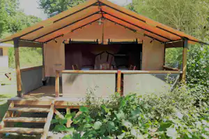 Lodge-Zelt mit Terrasse, Holzbank und Tisch, Lichterkette und Blick ins Innere, umgeben von Bäumen und Grün.
