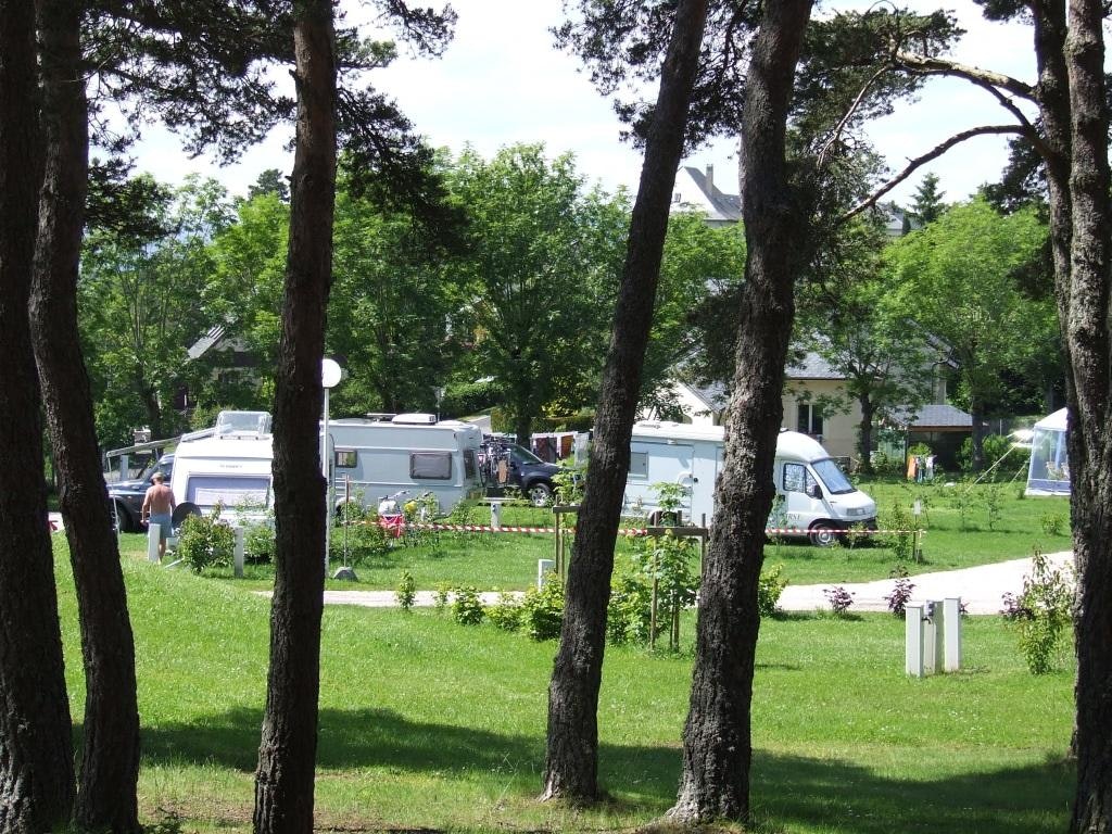 Wohnmobile und ein Auto stehen auf einem Campingplatz zwischen hohen Bäumen, mit gepflegten Wegen und Rasenflächen.