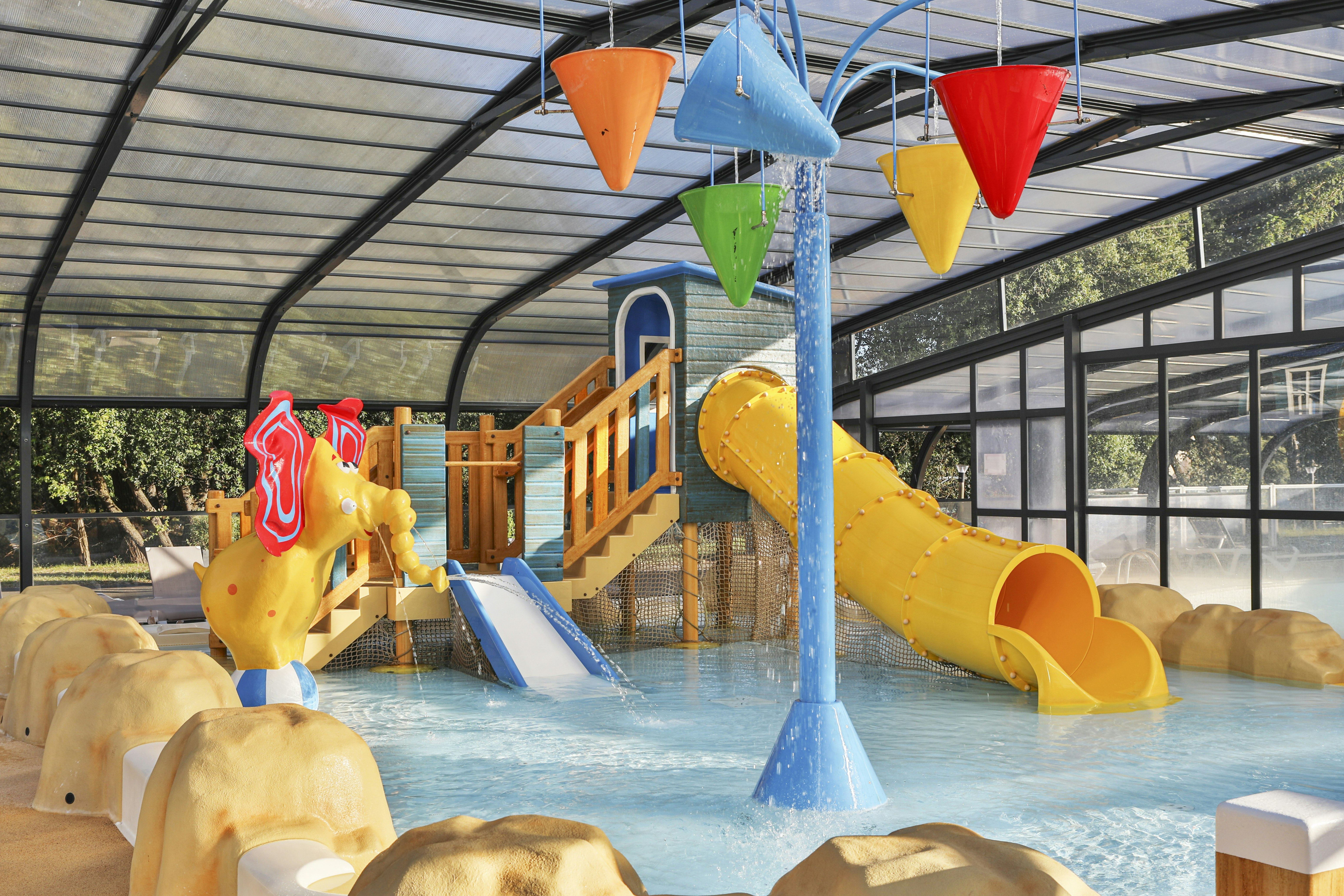 Innenbereich mit Kinder-Wasserrutschen und kleinem Pool, bunten Rutschen und Spielgeräten unter einem Glasdach.