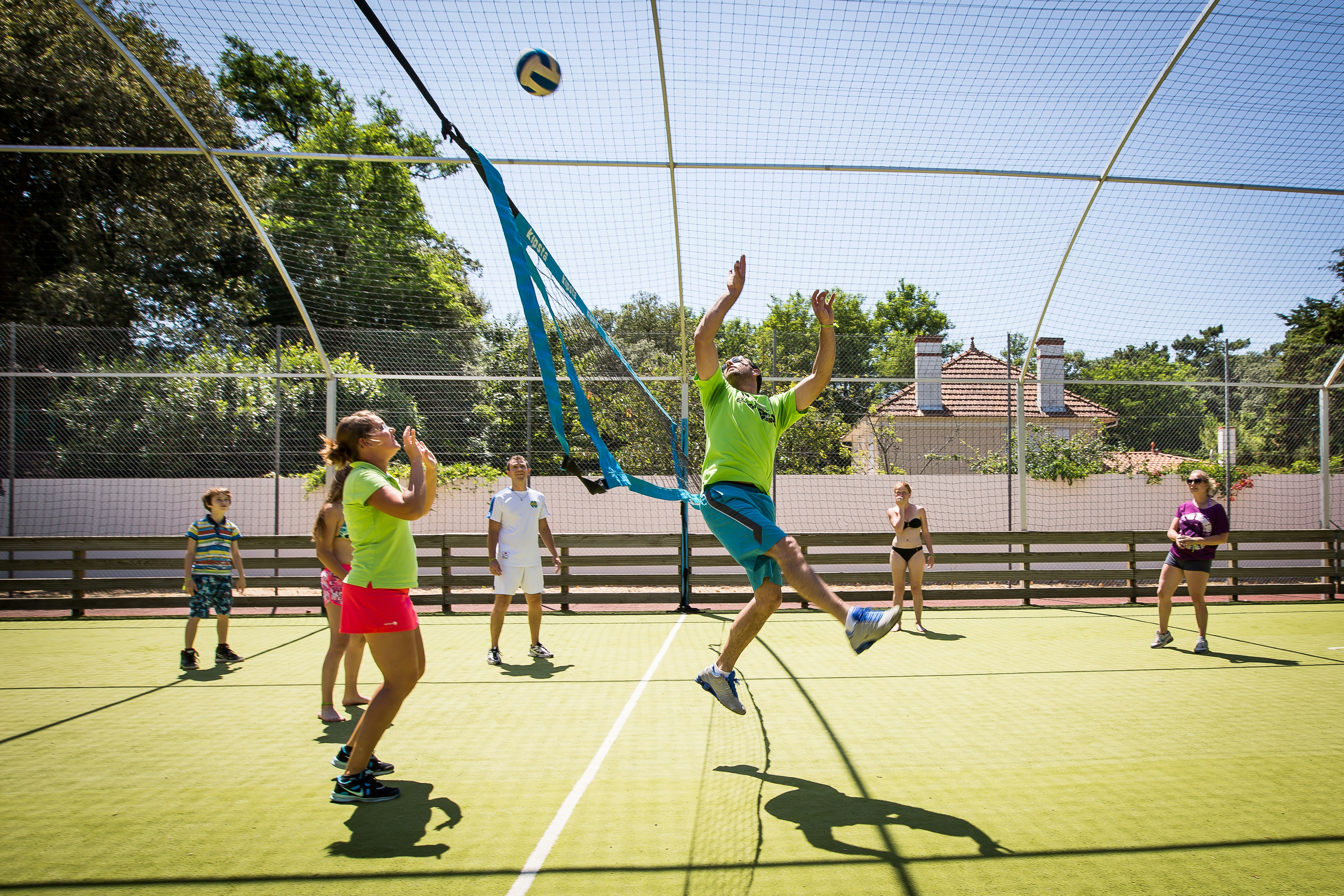 Kinder spielen auf einem Tennisplatz im Campingbereich, ein Kind springt mit Ball, im Hintergrund Zaun und Bäume.