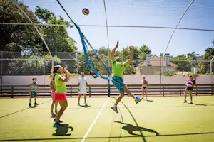 Kinder spielen auf einem Tennisplatz im Campingbereich, ein Kind springt mit Ball, im Hintergrund Zaun und Bäume.